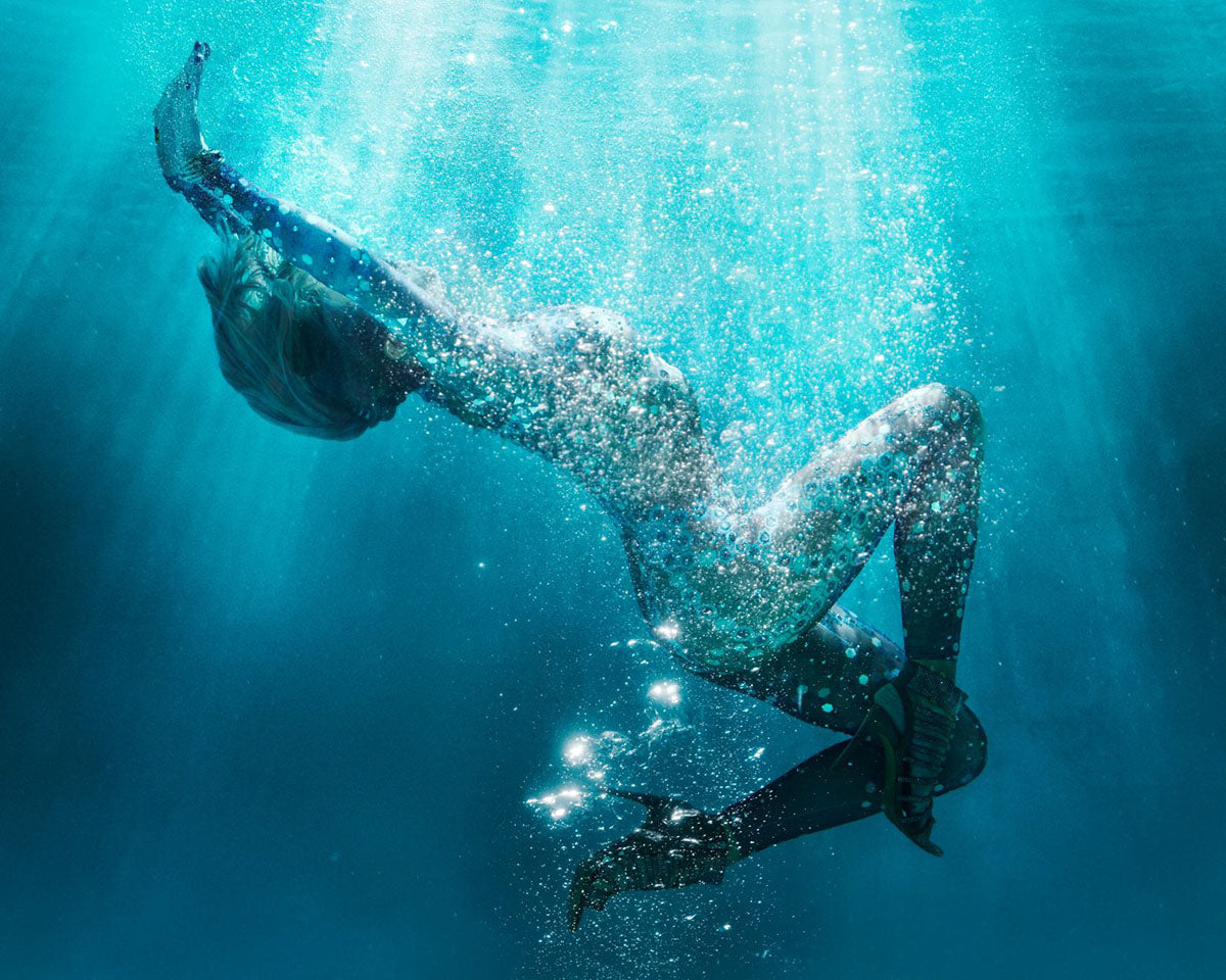 A person underwater in a dark swimsuit, captured mid-dive with bubbles and light rays filtering through the blue water.
