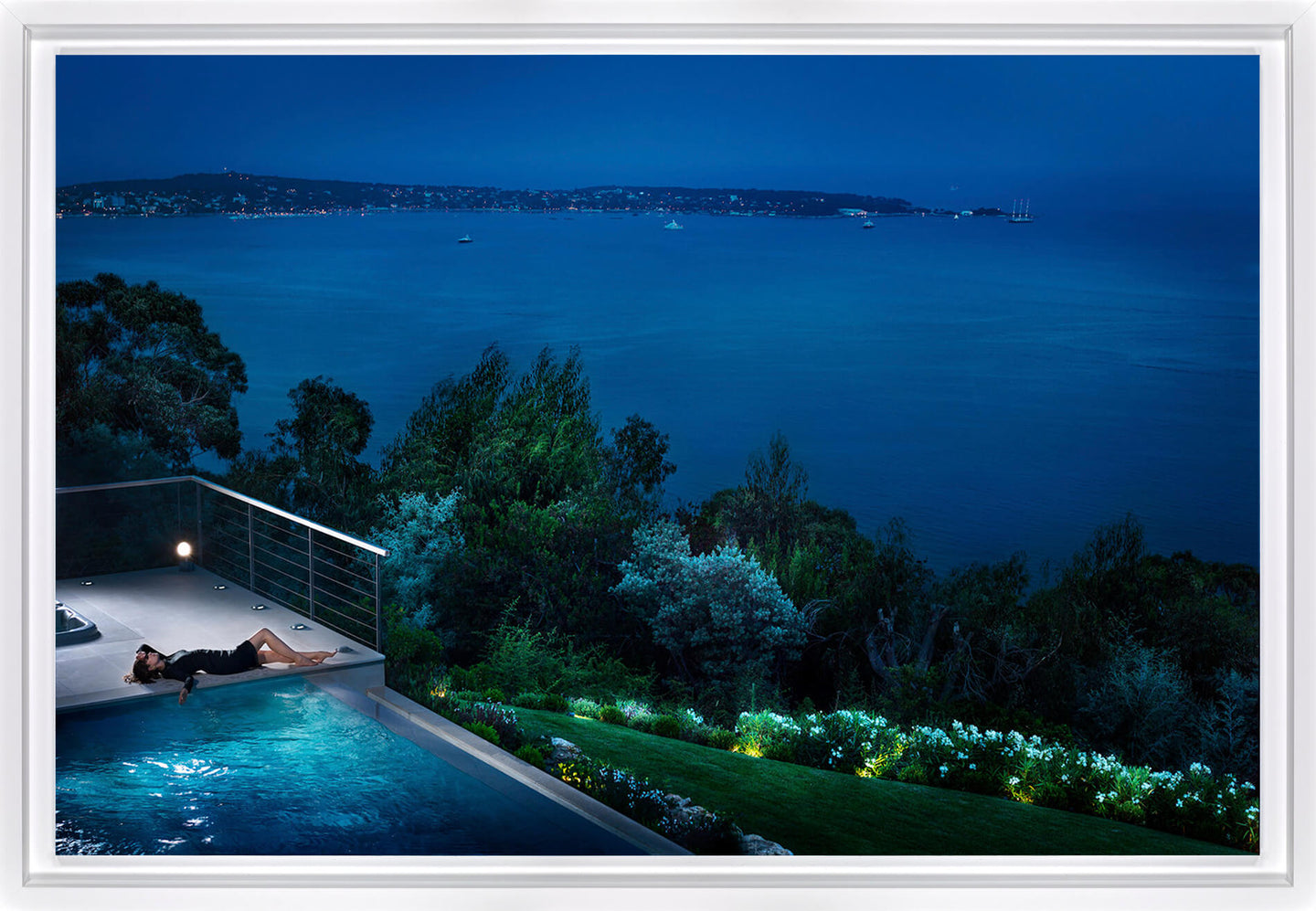 A framed art print depicting a luxurious coastal night scene with a person relaxing by an infinity pool overlooking a deep blue ocean and distant shoreline.