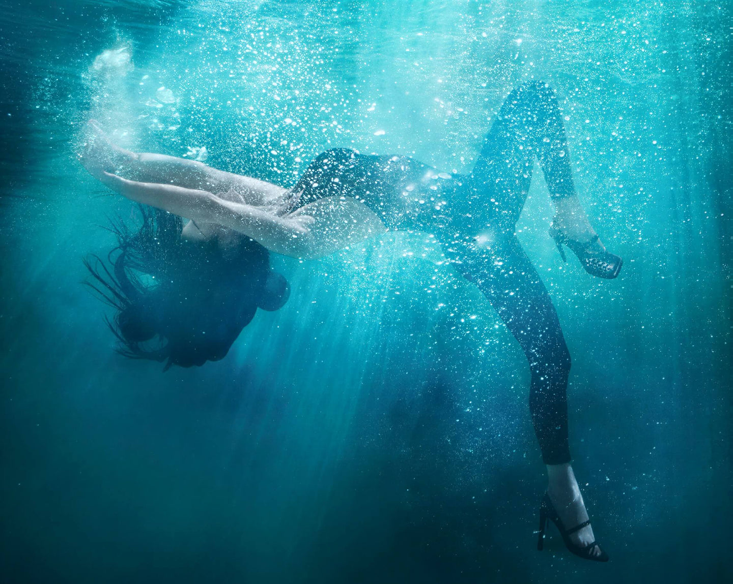 A woman swims underwater in a black swimsuit and black high heels, with bubbles and light rays filtering through the blue water.