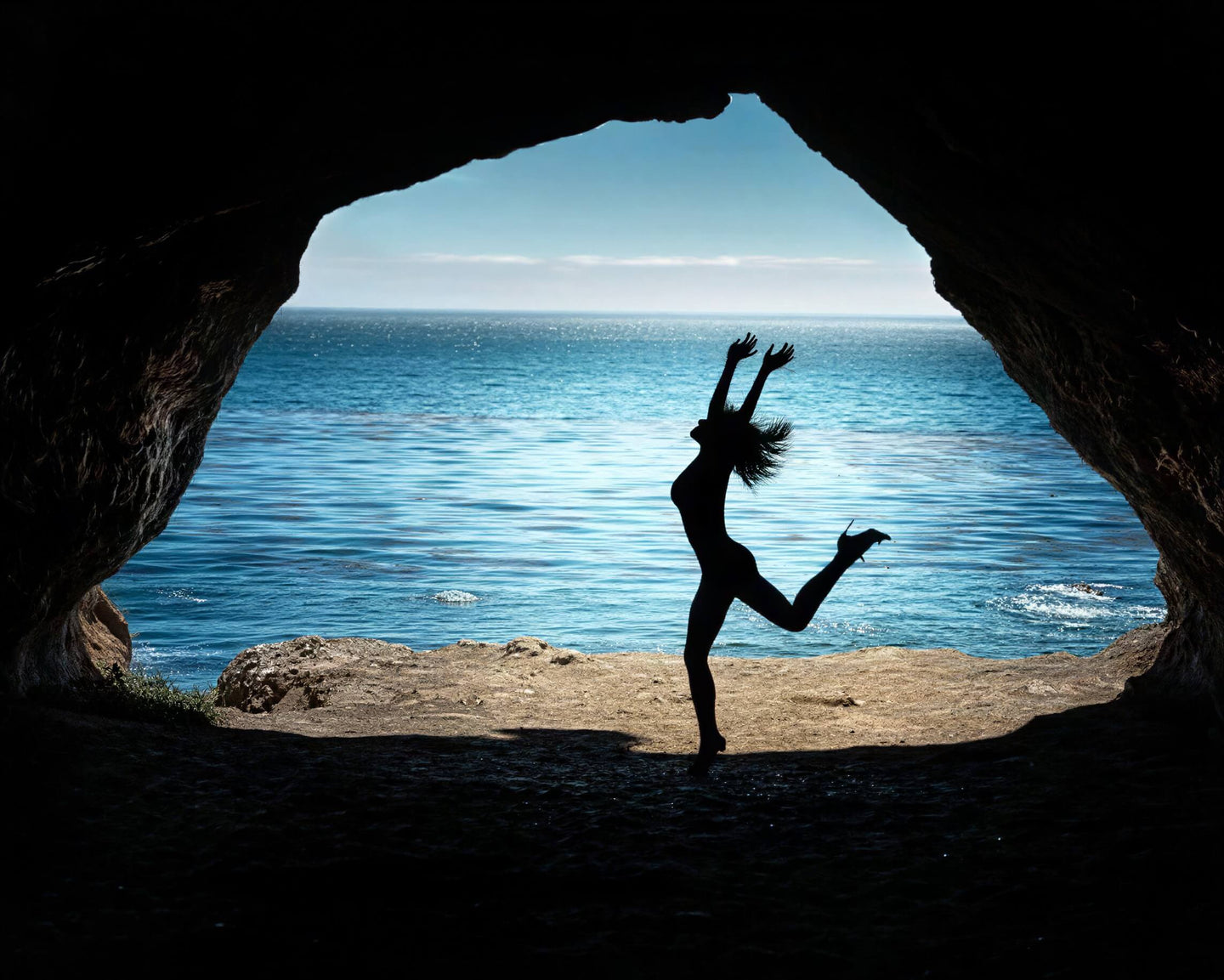 A silhouetted person jumping in front of a cave opening that frames a bright blue ocean scene, creating a dramatic high-contrast silhouette effect.