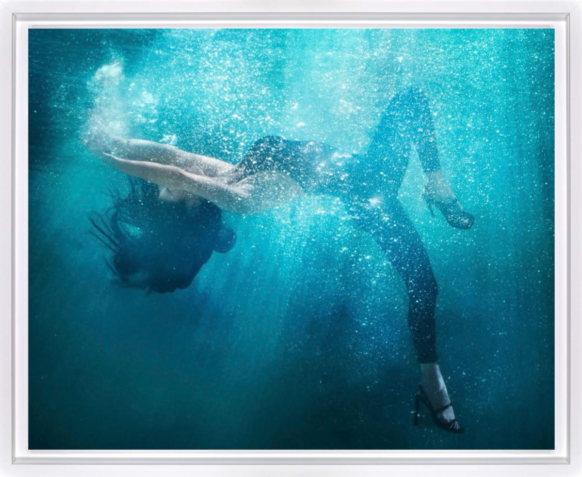 Underwater art print featuring a woman in a black swimsuit and heels swimming horizontally through turquoise blue water with rising bubbles and light rays filtering down, presented in a white frame.