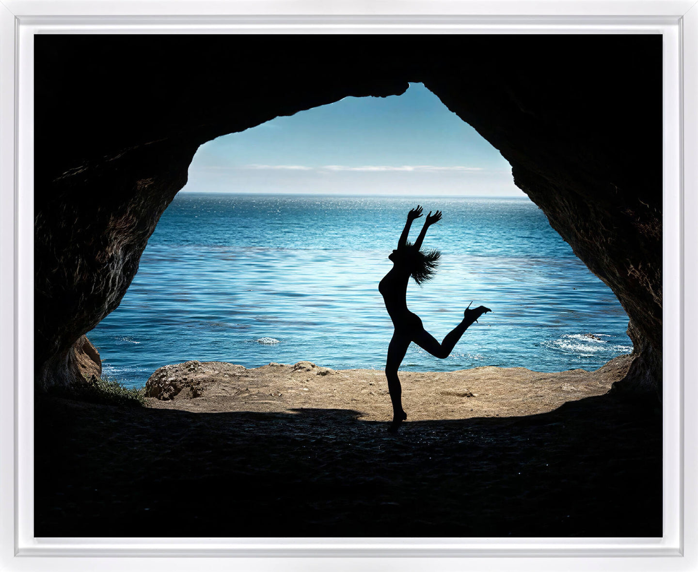 A framed photographic print showing a silhouetted dancer jumping in front of a cave opening that reveals a bright blue ocean and sky.