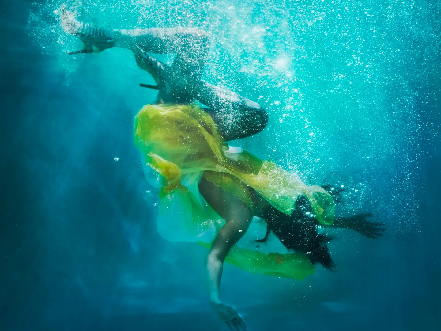 Underwater photograph of a person floating upside down in clear blue water with rising bubbles, wearing a flowing translucent yellow-green dress or fabric.