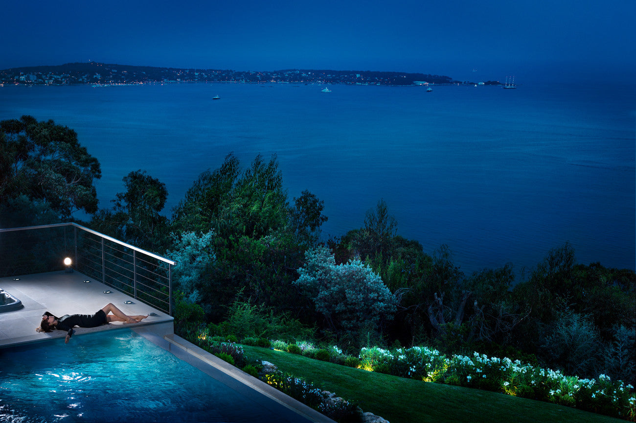 A person relaxes on a poolside deck overlooking a dark blue ocean at night, with illuminated landscaping, trees, and a distant shoreline visible.